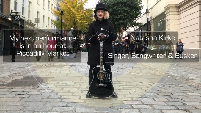 A busker with a guitar on a cobbled street wearing a Hex Beacon, labelled Singer, Songwriter and Busker